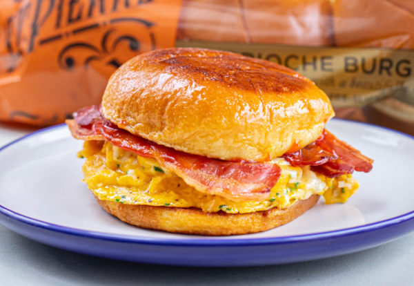 A photo of a Brioche French Toast Cheese and Bacon Roll recipe on a white and blue plate with St Pierre Brioche Burger Buns in the background