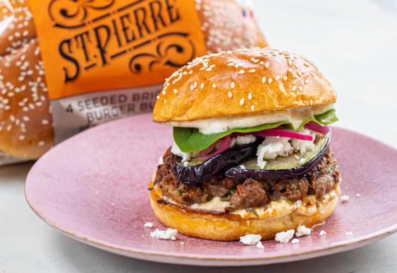 Photo credit: MOB Kitchen A Cumin Lamb and Aubergine BBQ Burger on a pink plate, placed on a white table with St Pierre burger buns in the background