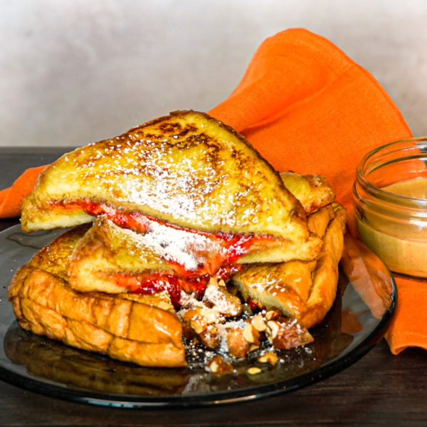 A photo of a peanut butter and strawberry jam French toast sandwich on a glass plate with an orange napkin and jar of peanut butter next to it
