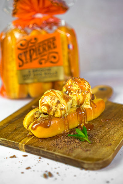 A photo of a brioche hot dog roll on a wooden board filled with scoops of vanilla ice cream and caramel sauce